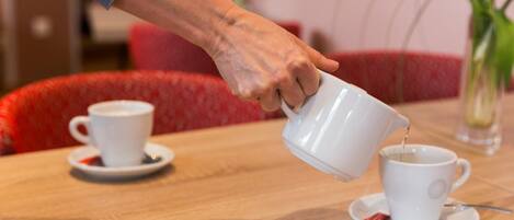 Een hand schenkt koffie in een kopje op een houten tafel, met nog een kop en schotel op de achtergrond.