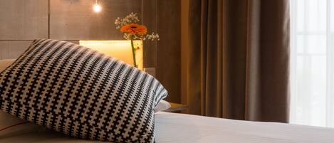 A hotel room with a bed, a patterned pillow, a wall-mounted lamp, a vase with flowers, and a window with curtains.