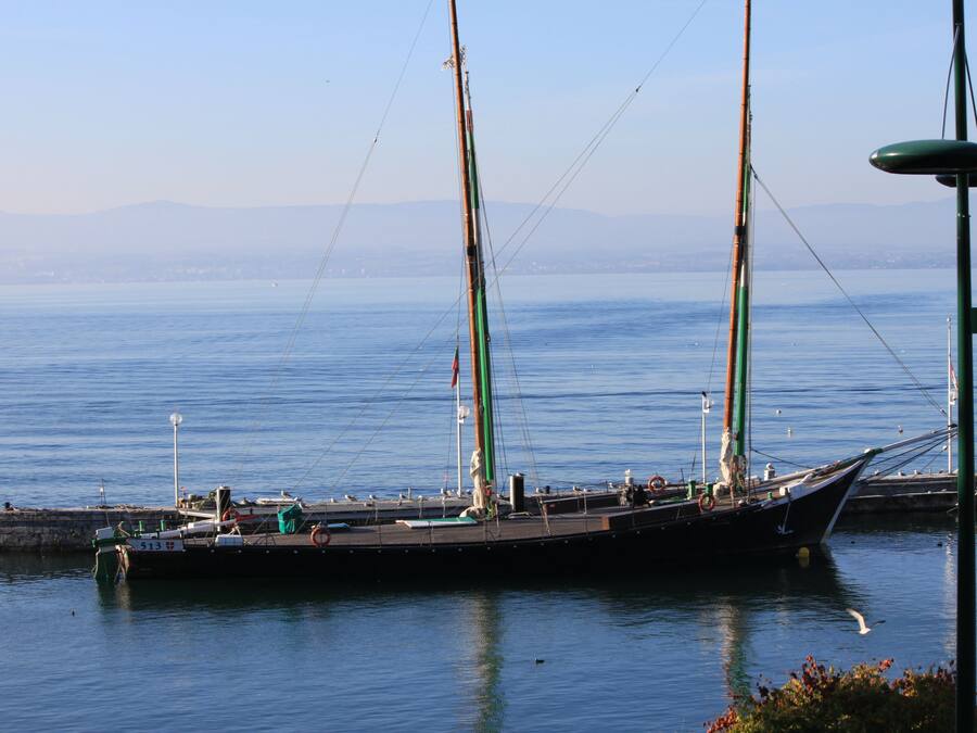 A large sailing ship docked at a pier with calm water and distant mountains in the background.