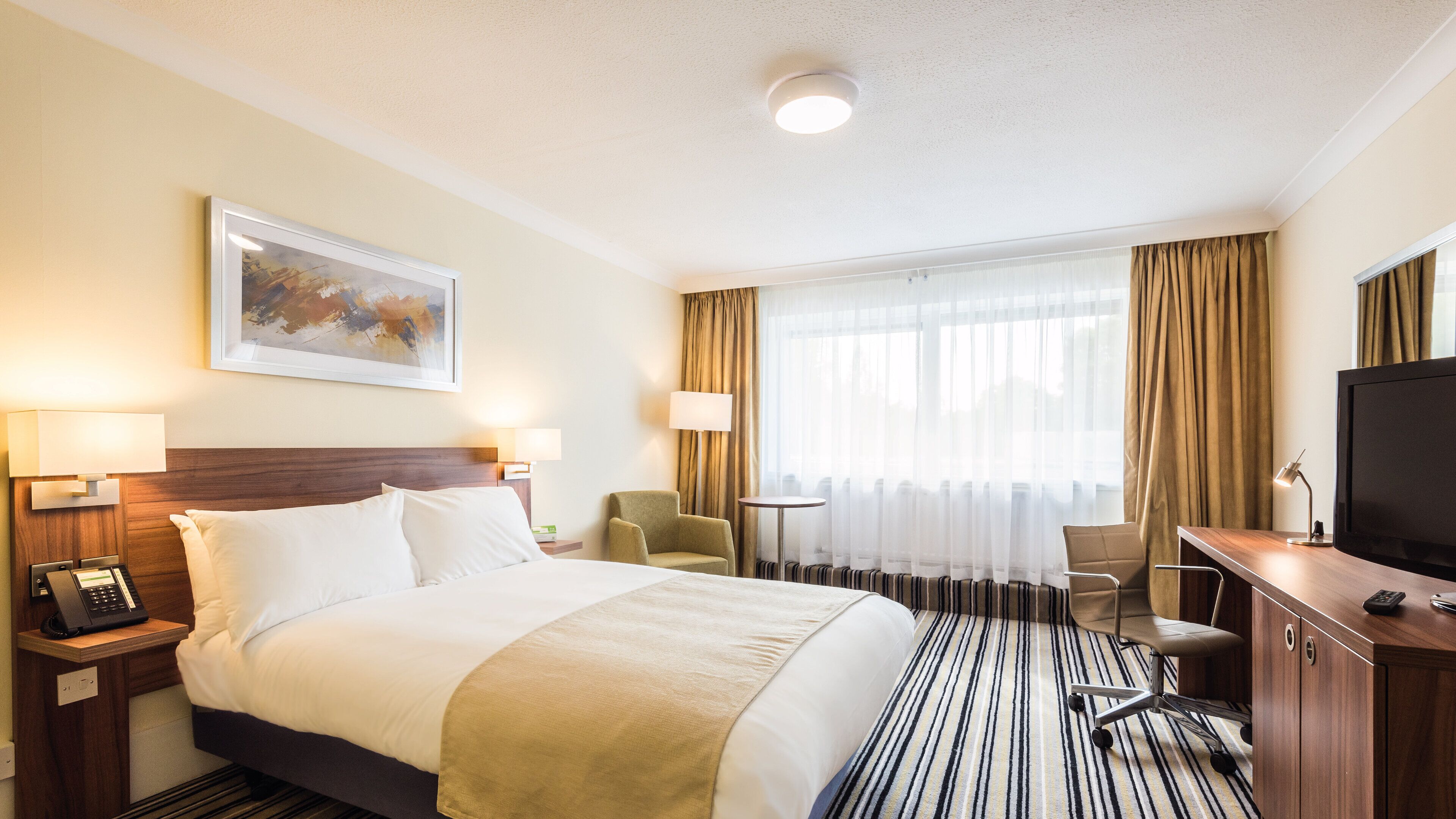 A hotel room with a bed, two bedside lamps, a desk with a TV, a chair, a small table, and a painting on the wall.