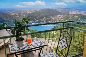 Un balcón con una planta en maceta y un vaso de jugo de naranja, con vistas panorámicas a un lago y colinas circundantes.