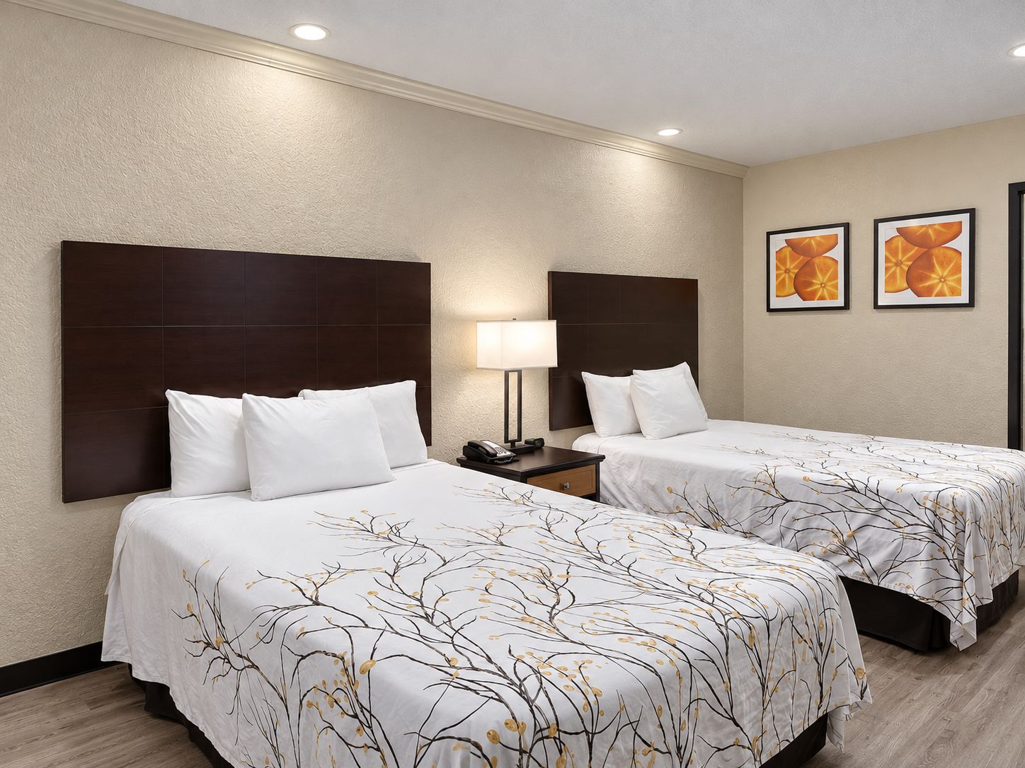 Habitación de hotel con dos camas, cada una con cabecero oscuro y ropa de cama blanca. Hay una mesita de noche con lámpara y teléfono. Las paredes son color beige y hay dos cuadros enmarcados.