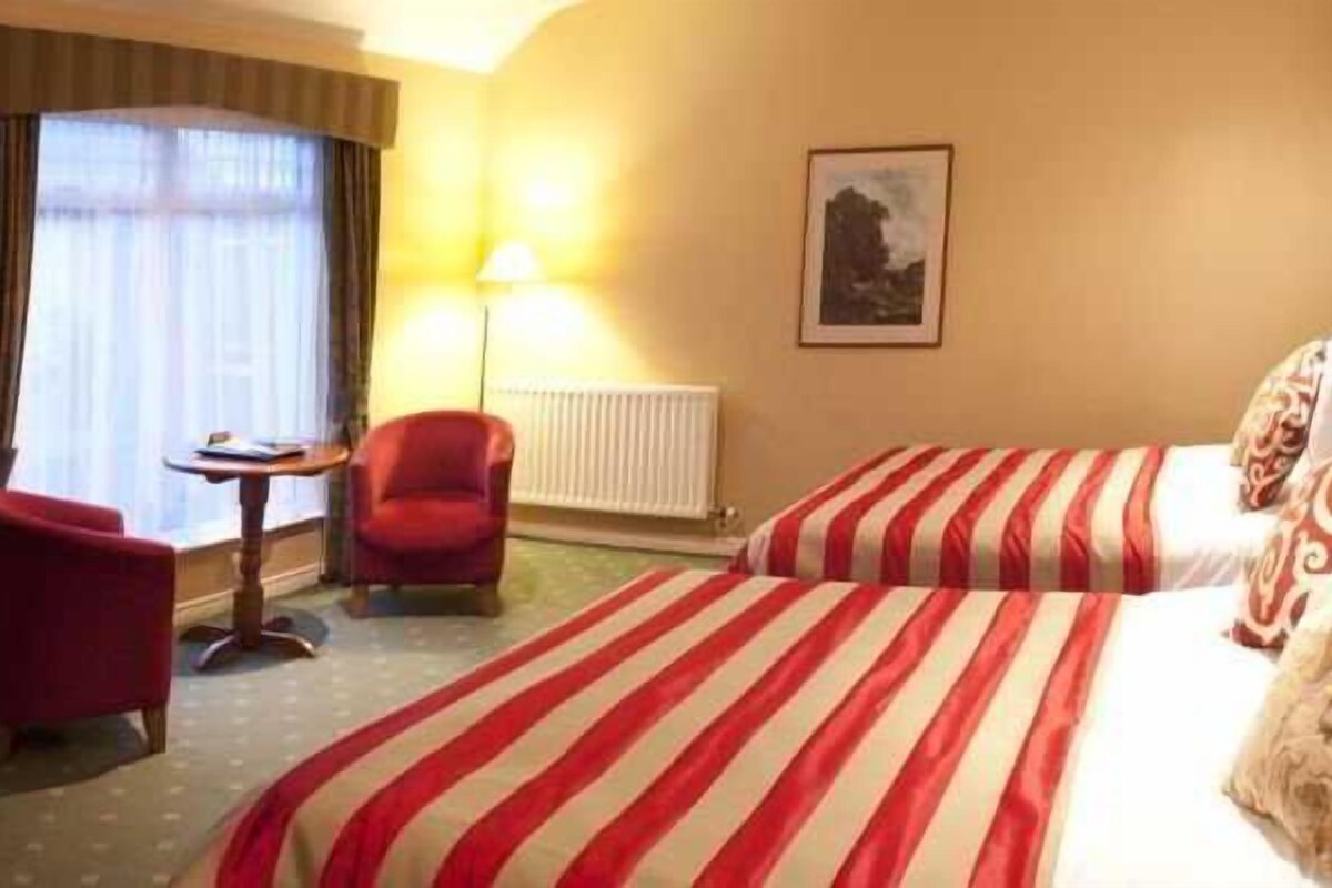 A hotel room with two beds, a red armchair, a small table, and a lamp.