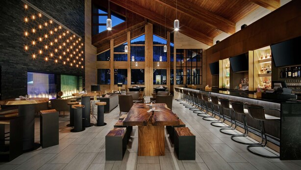 A spacious bar area with high wooden ceilings, a long counter with stools, and a large wooden table in the center. - Highline Vail - a DoubleTree by Hilton (Vail)