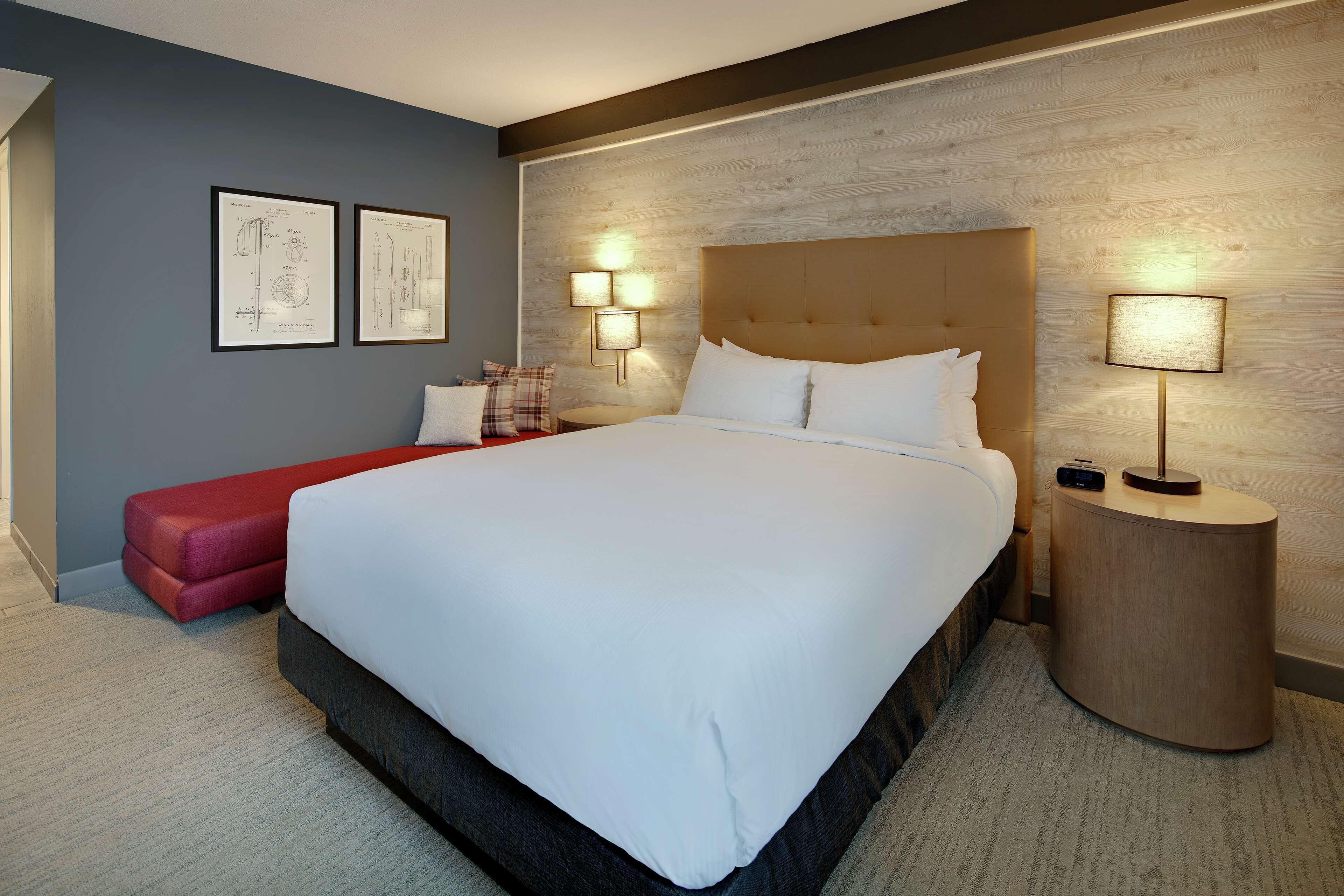 A modern hotel room with a large bed, a red bench, two wall-mounted lamps, and framed blueprints on the wall.