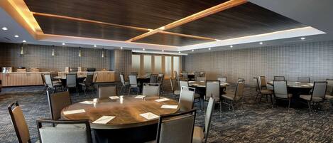 A conference room with a large round table surrounded by chairs, a kitchen area in the background, and a patterned carpet.