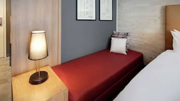 Une chambre d’hôtel avec un lit simple, une table de chevet avec une lampe, une couverture rouge et deux plans encadrés accrochés au mur.