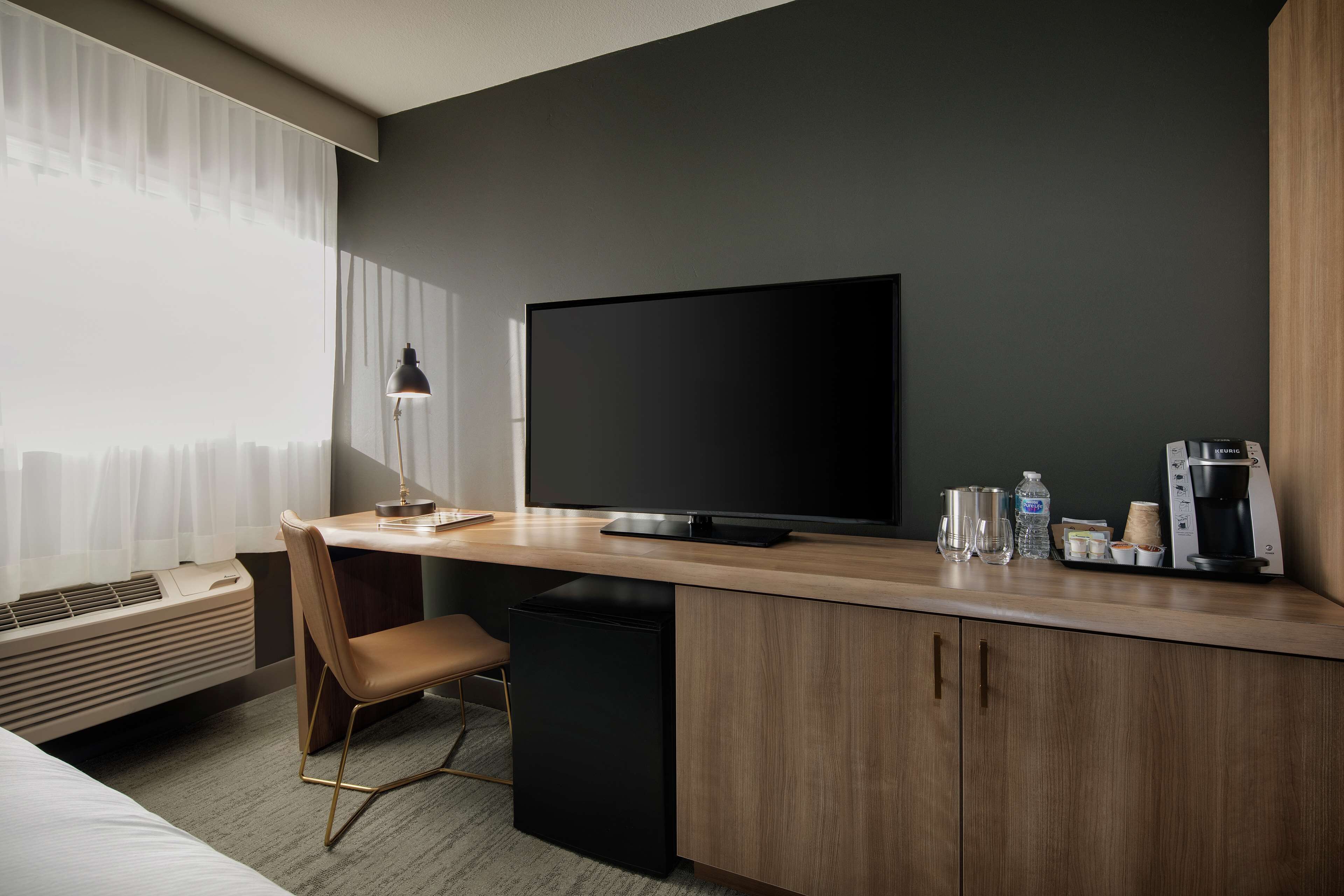 Una habitación de hotel moderna con un escritorio de madera, un televisor de pantalla plana, una silla, un pequeño cubo de basura negro y una cafetera.