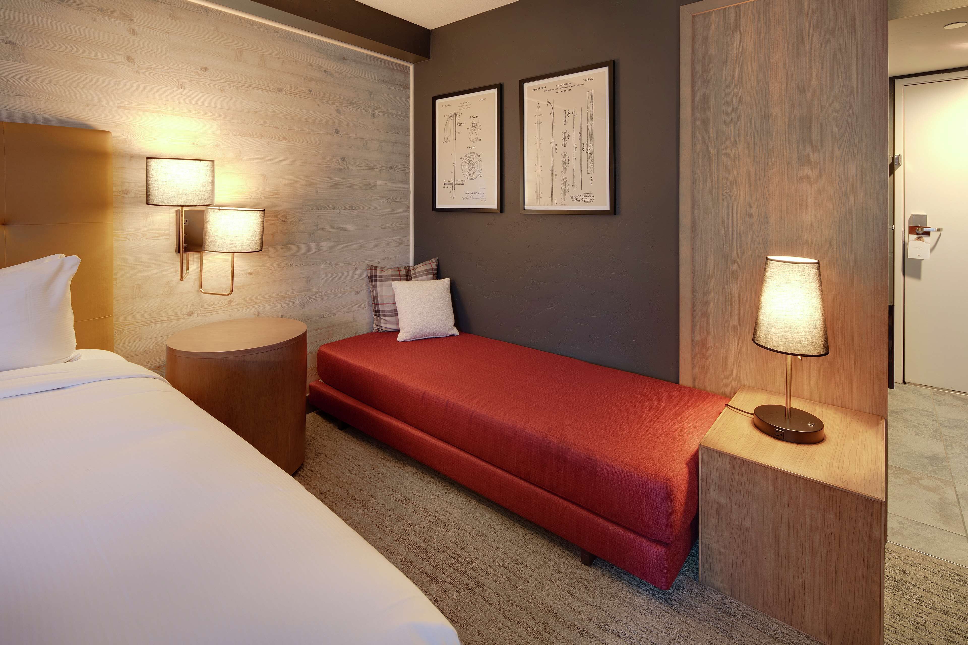 Hotellihuone, jossa on sänky, punainen sohva, pyöreä puinen sivupöytä, lamppu ja kaksi kehystettyä piirustusta seinällä.