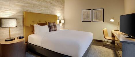 A hotel room with a bed, bedside lamps, a desk with a television, and framed architectural drawings on the wall.