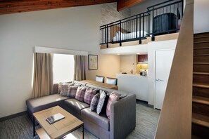A modern living room with a sofa, coffee table, and a dining area. A staircase leads to an upper level with a balcony.