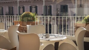 A balcony with white chairs and tables, potted plants, and a view of buildings through a glass railing.