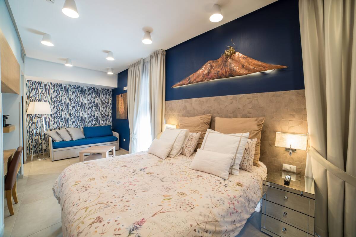 A bedroom with a large bed, a nightstand, a lamp, a sofa, and a patterned wall.