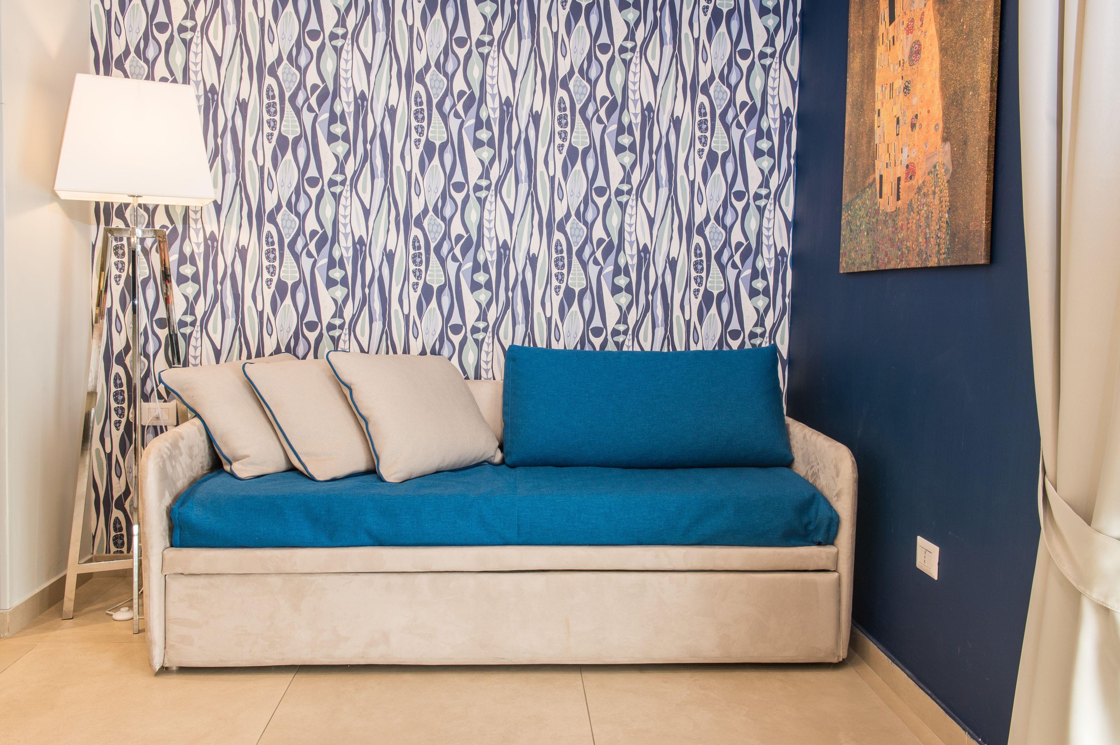 A sofa with a blue cover and beige cushions, a floor lamp, and a patterned wallpaper.