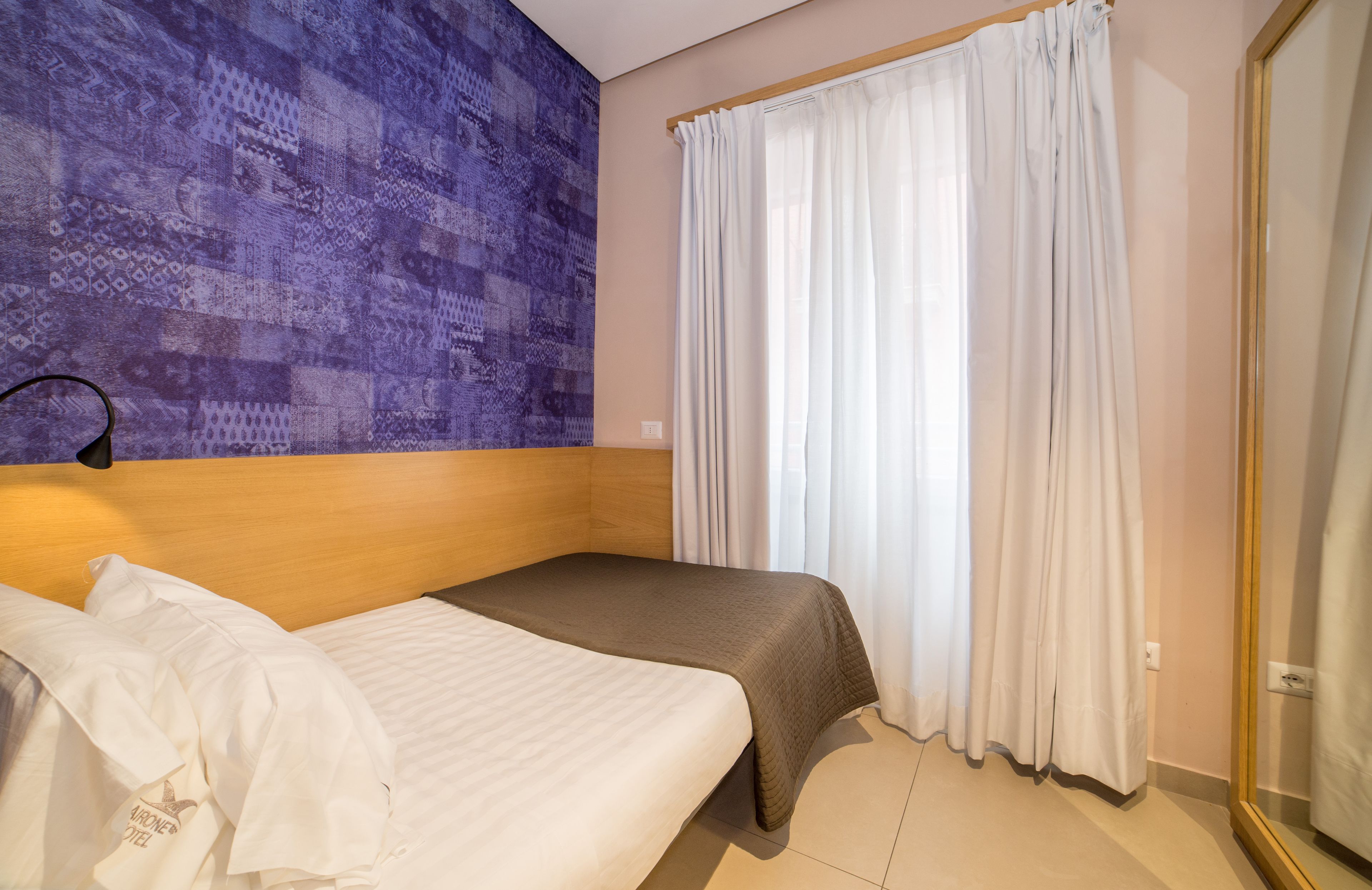 A hotel room with a bed, a headboard, a wall with a patterned design, and a window with curtains.