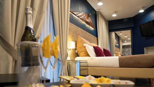 A hotel room with a bed, a dining table with fruit, a bottle of wine, and a TV on the wall.