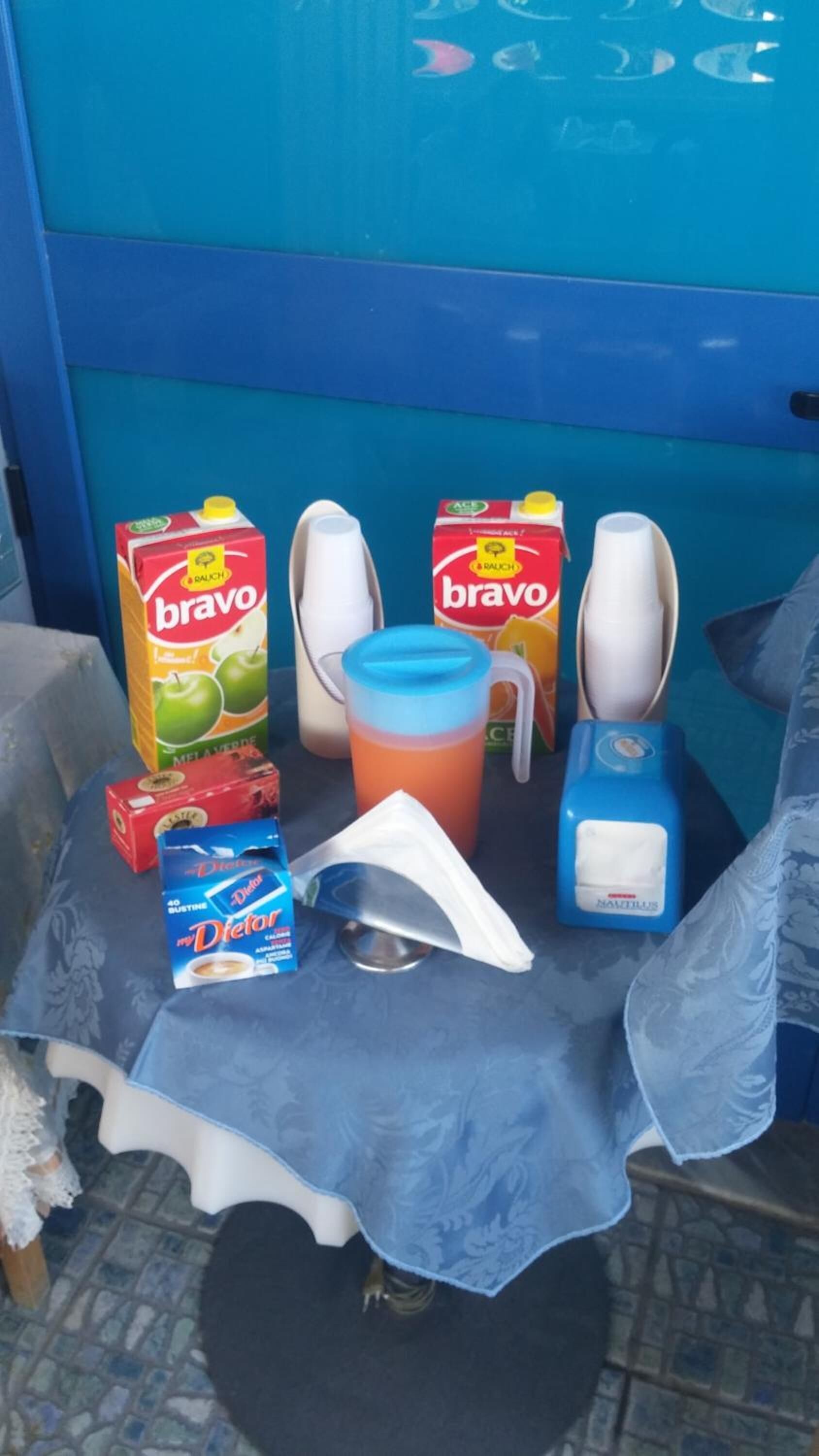 Ein Tisch mit einer blauen Tischdecke, zwei Kartons 'bravo'-Saft, eine weiße Tasse, eine blaue Wasserflasche, ein weißes Servietttuch und eine blaue Schachtel 'Dietor'-Energieriegel.