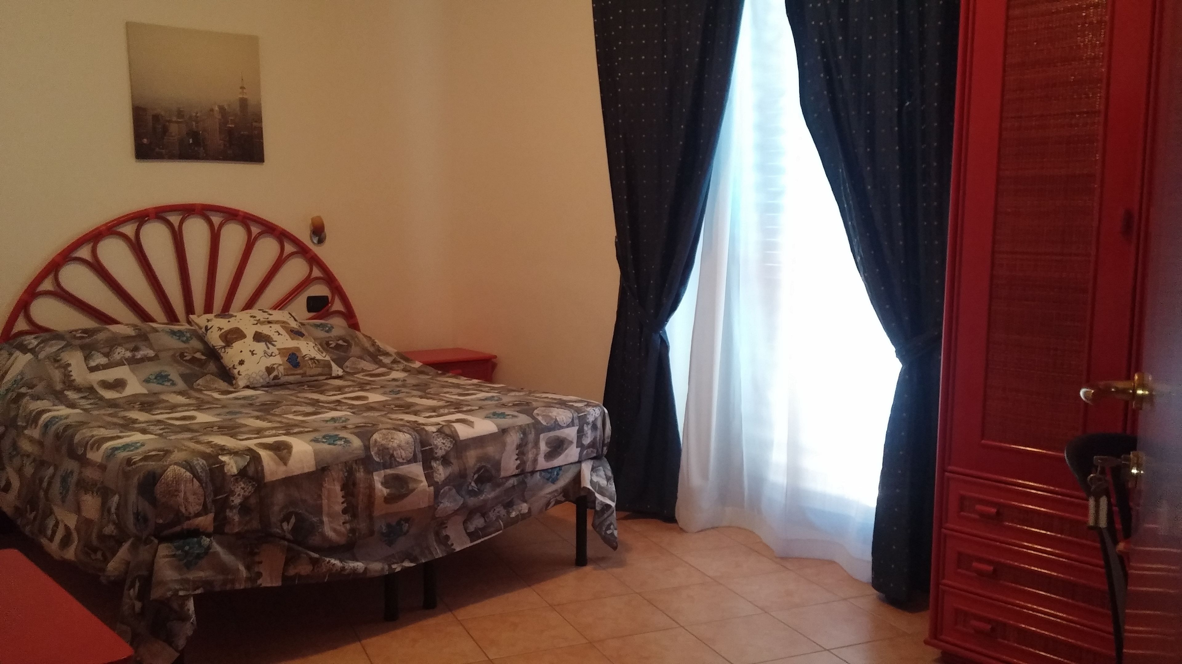 A bedroom with a patterned bedspread, a red metal bed frame, a small bedside table, a red wardrobe, and a window with dark curtains.