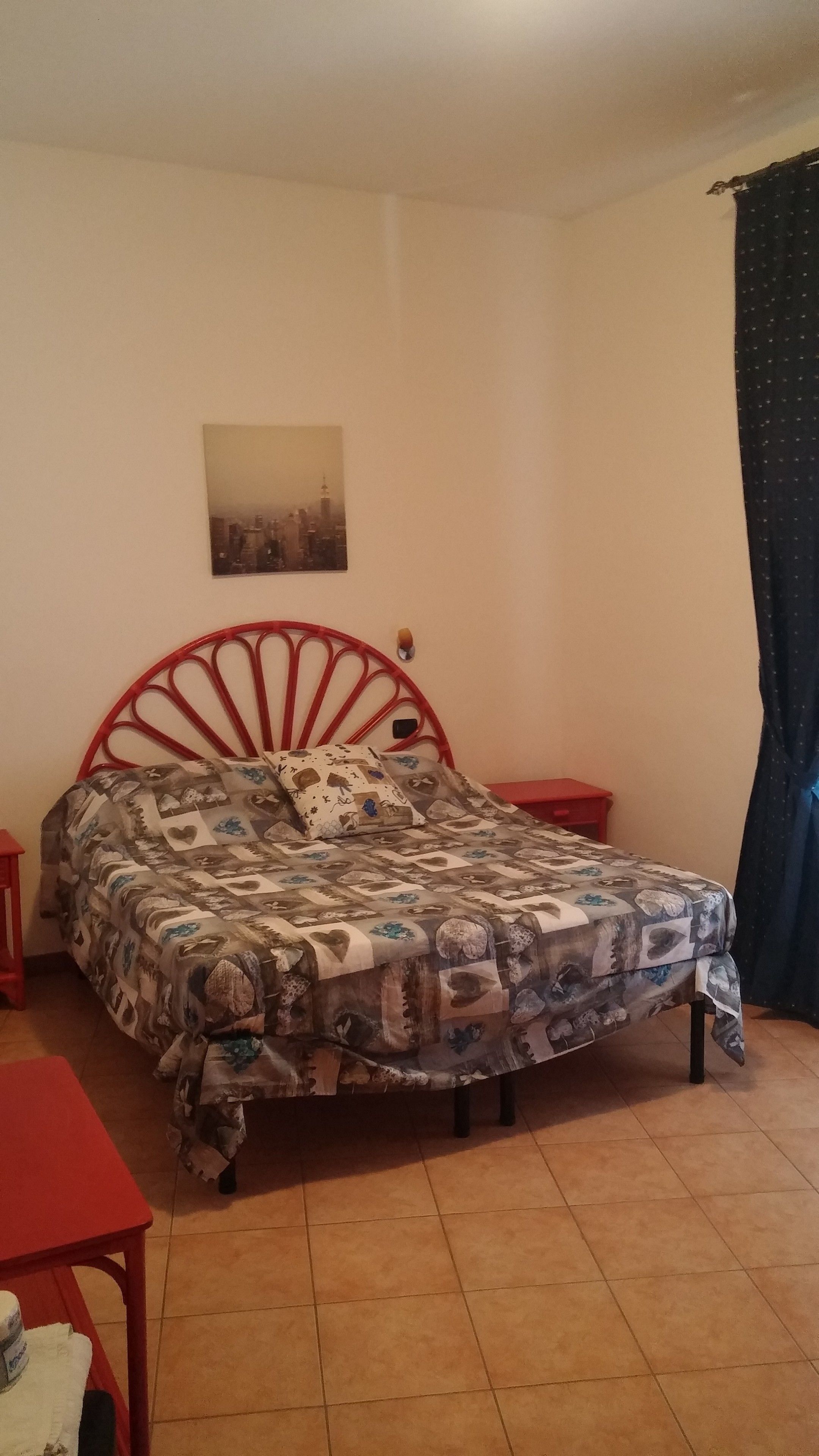 Un dormitorio con estructura de cama metálica roja, colcha estampada, mesita de noche pequeña, una silla roja y una mesa roja.