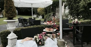 Un área de comedor al aire libre con mesas dispuestas para una comida, rodeada de follaje y una sombrilla blanca.