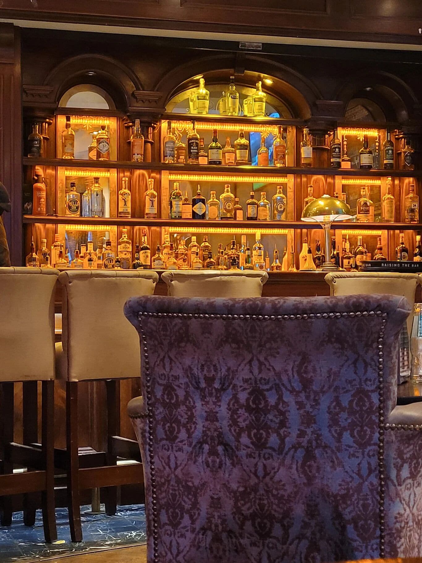 A bar area with multiple shelves of liquor bottles, bar stools, and a patterned chair in the foreground.