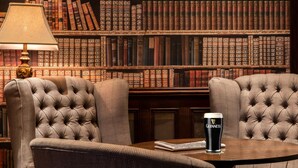 A library setting with bookshelves, two tufted armchairs, a table with a glass of Guinness beer, and a lamp. - Headfort Arms Hotel (Kells)