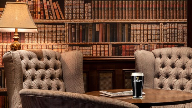 A library setting with bookshelves, two tufted armchairs, a table with a glass of Guinness beer, and a lamp. - Headfort Arms Hotel (Kells)