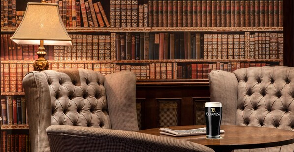 A library setting with bookshelves, two tufted armchairs, a table with a glass of Guinness beer, and a lamp.
