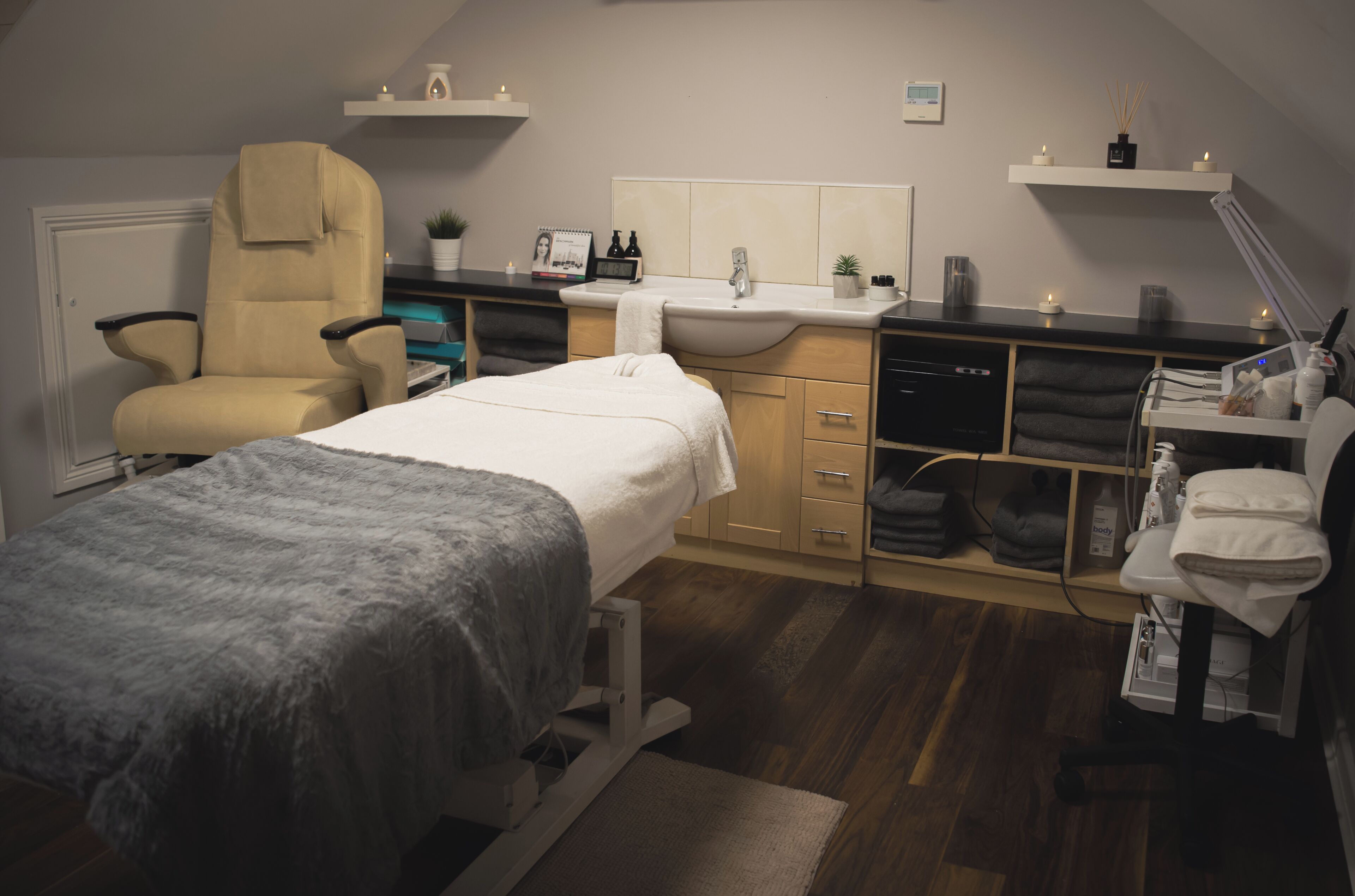 A room with a bed, a chair, a desk with a computer, a sink, and shelves with various items.