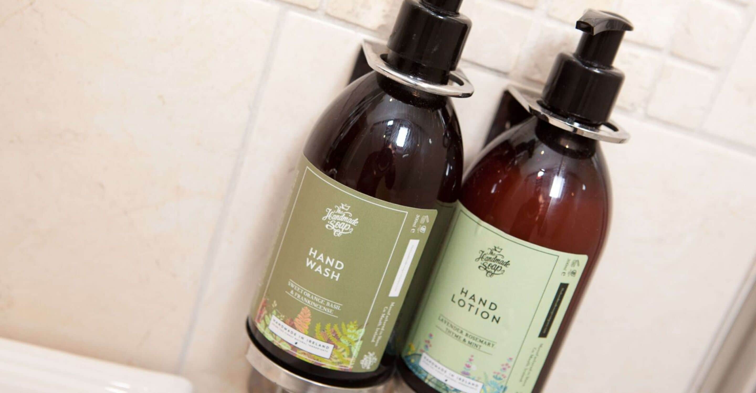 Dos botellas de jabón artesanal, una con la etiqueta 'Hand Wash' y la otra 'Hand Lotion', sobre una superficie de azulejos.