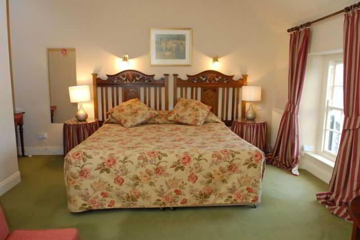 A bedroom with a floral bedspread, two bedside lamps, a headboard with a picture, and striped curtains.