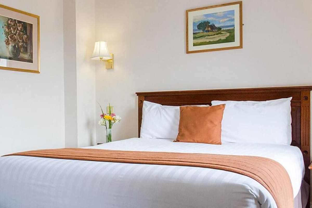 A neatly made bed with white and brown bedding, a wooden headboard, and a nightstand with a vase of flowers.