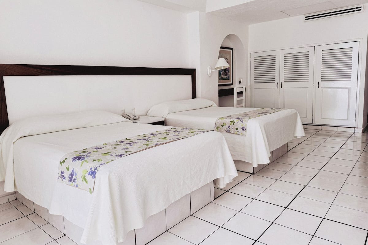 Habitación de hotel con dos camas, ropa blanca de cama, cabecero de madera, suelo de baldosas y un armario blanco con puertas persianas.