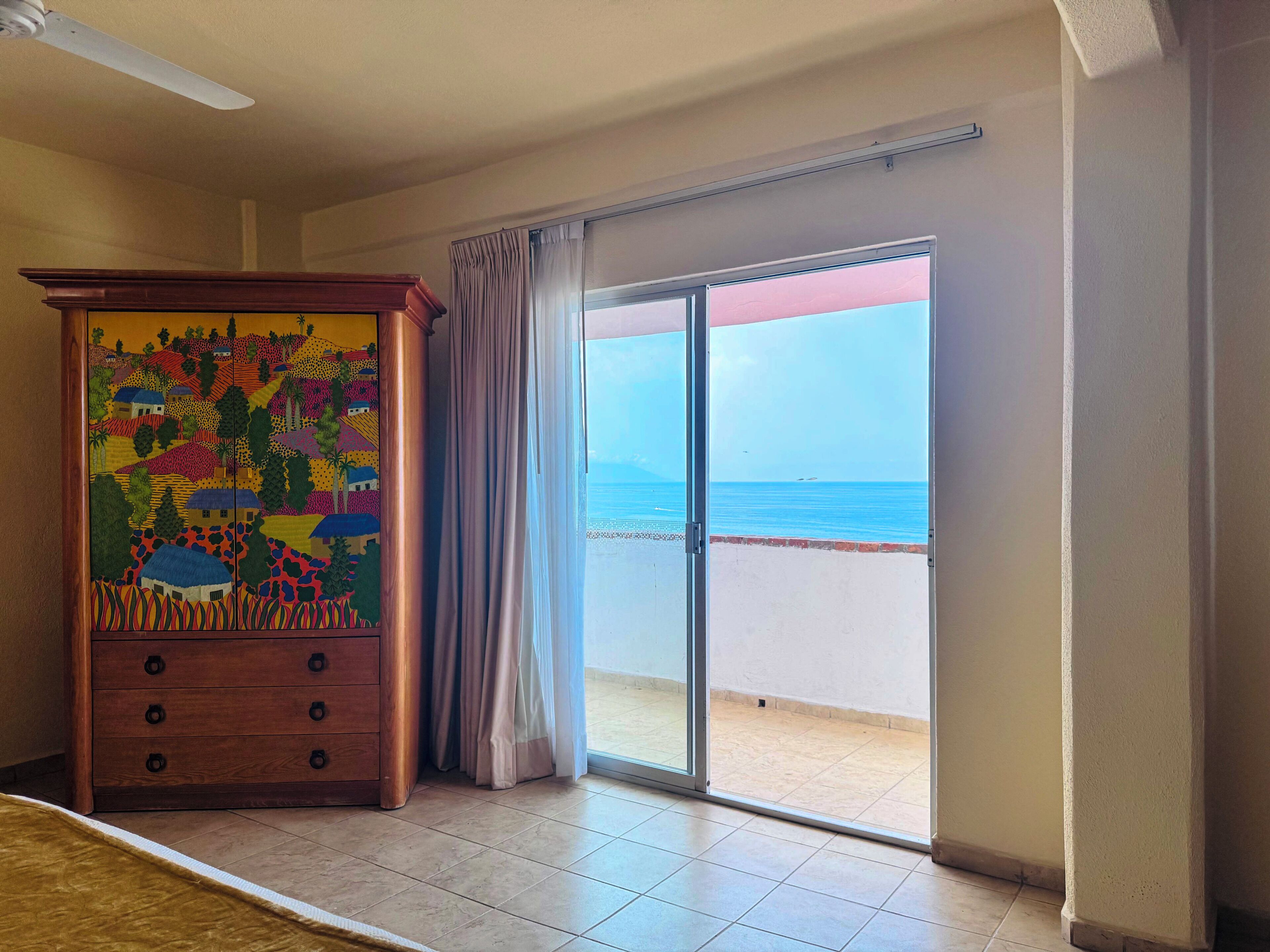 Une pièce dotée d’une grande commode en bois et d’une porte-fenêtre coulissante donnant sur un balcon avec vue sur la mer.