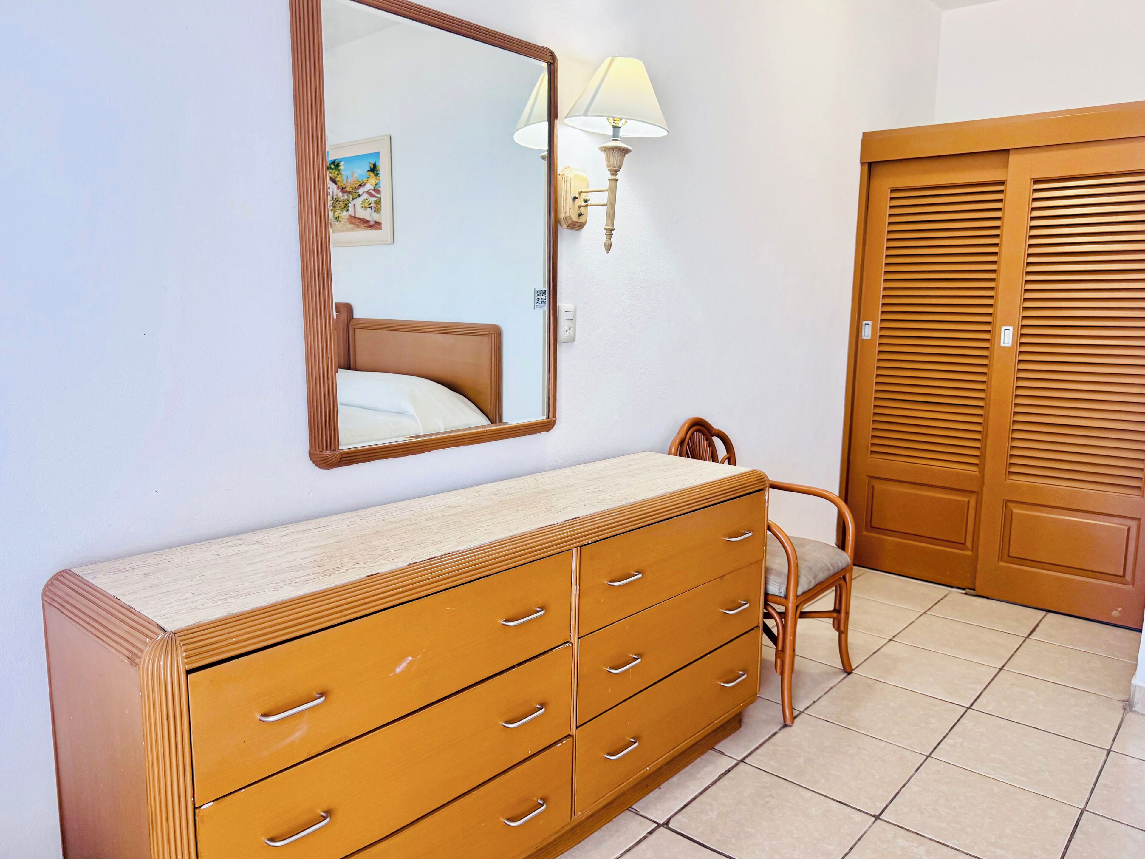A bedroom with a wooden dresser, a chair, a bed, a mirror, and a wall-mounted lamp.