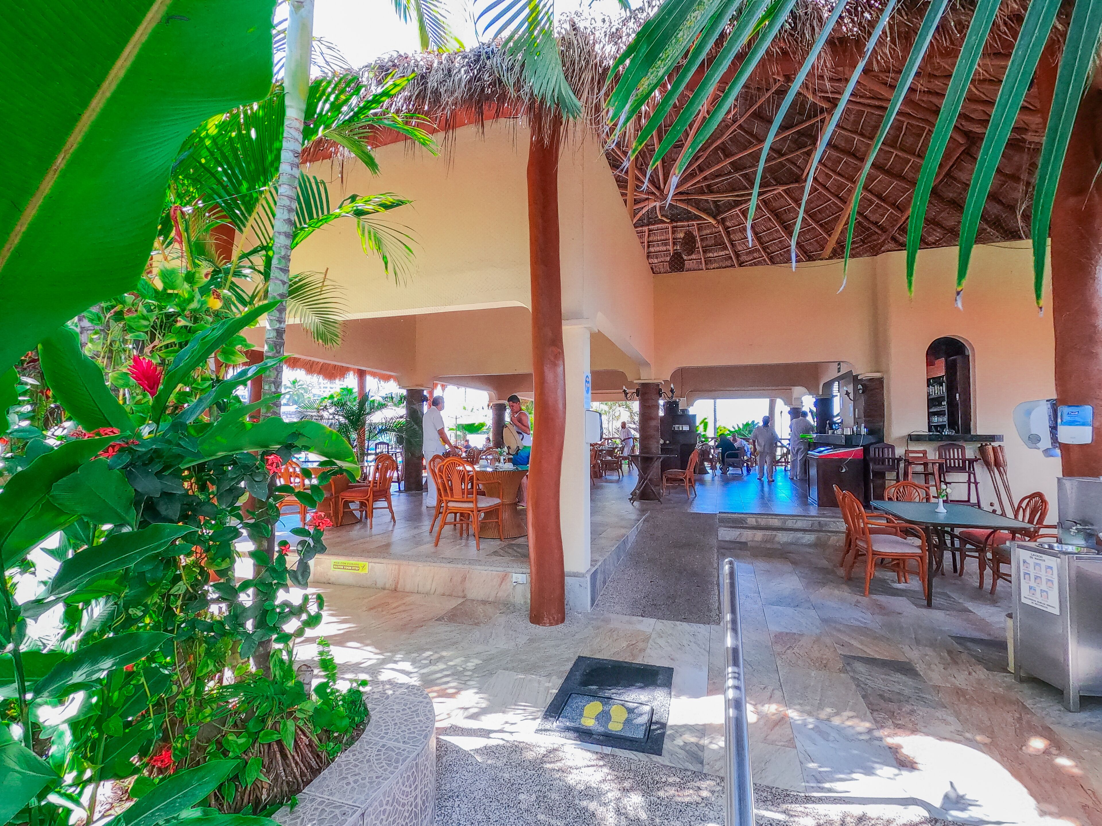 A spacious outdoor area with a pool, dining tables, and chairs, surrounded by lush greenery and palm trees.