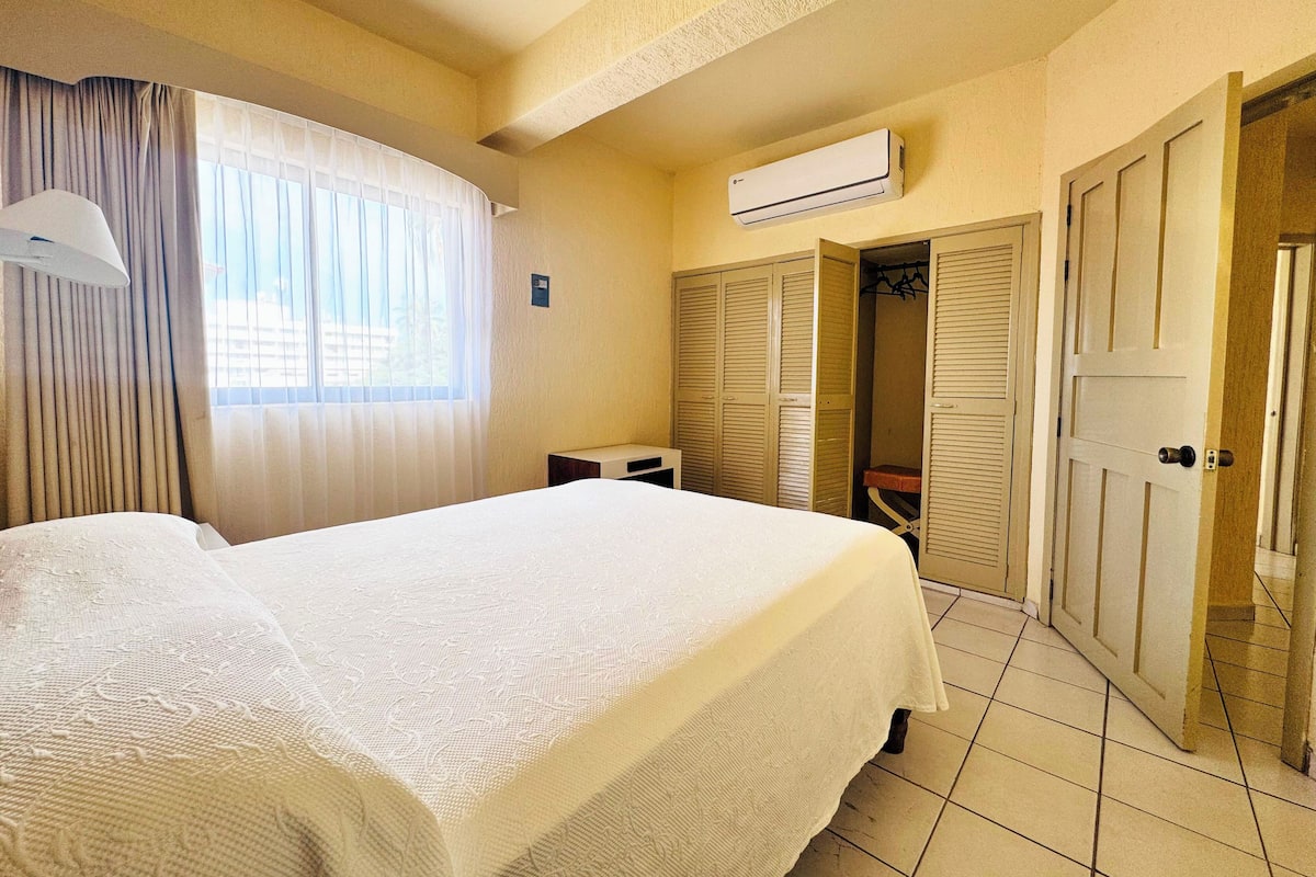Habitación de hotel con cama, ventana con cortinas, equipo de aire acondicionado, armario y puerta que conduce a otra habitación.
