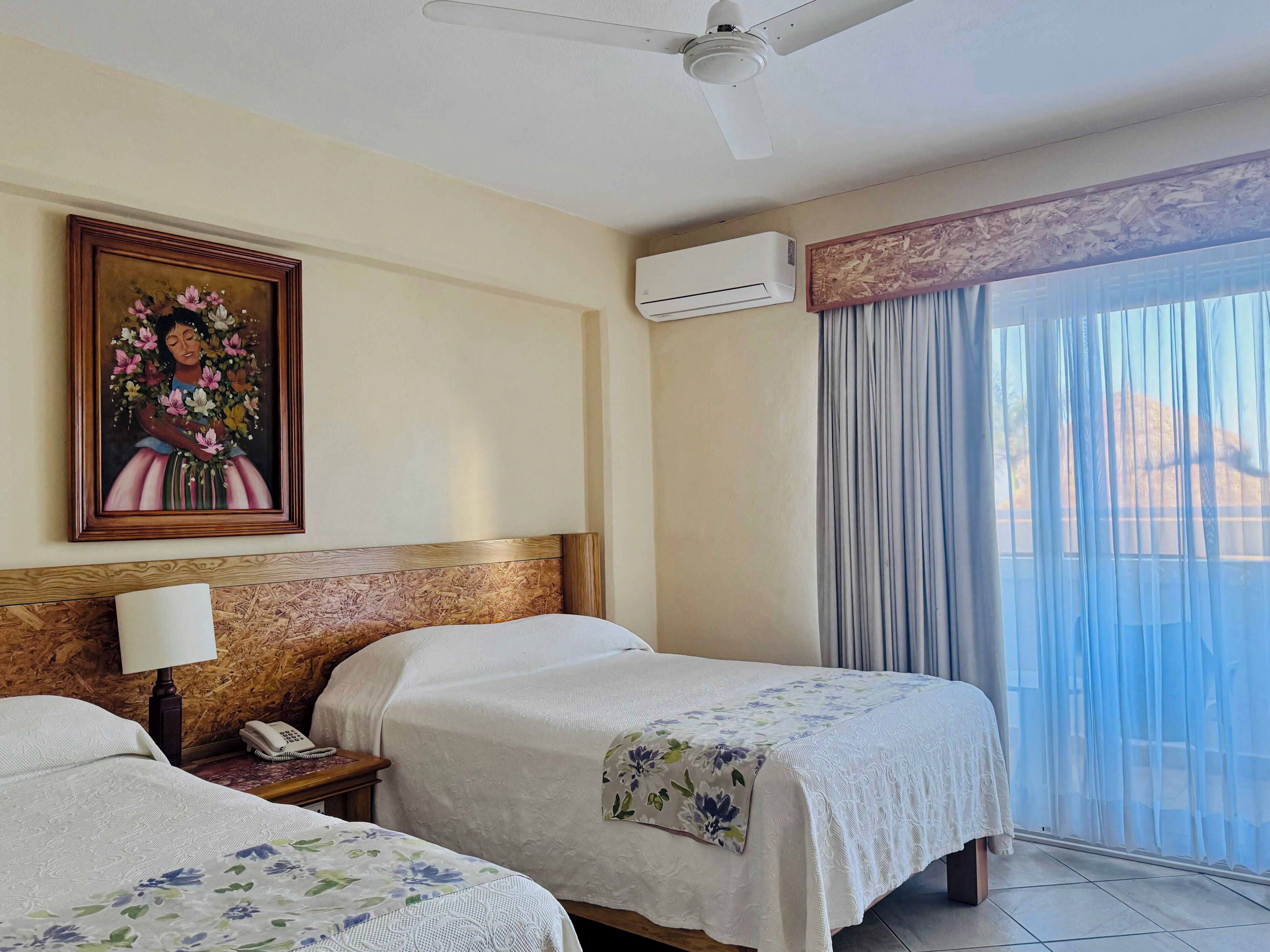 A hotel room with two beds, a ceiling fan, an air conditioning unit, a painting on the wall, and a view through sheer curtains.