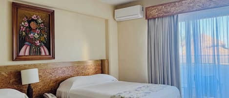 A hotel room with two beds, a ceiling fan, an air conditioning unit, a painting on the wall, and a view through sheer curtains.