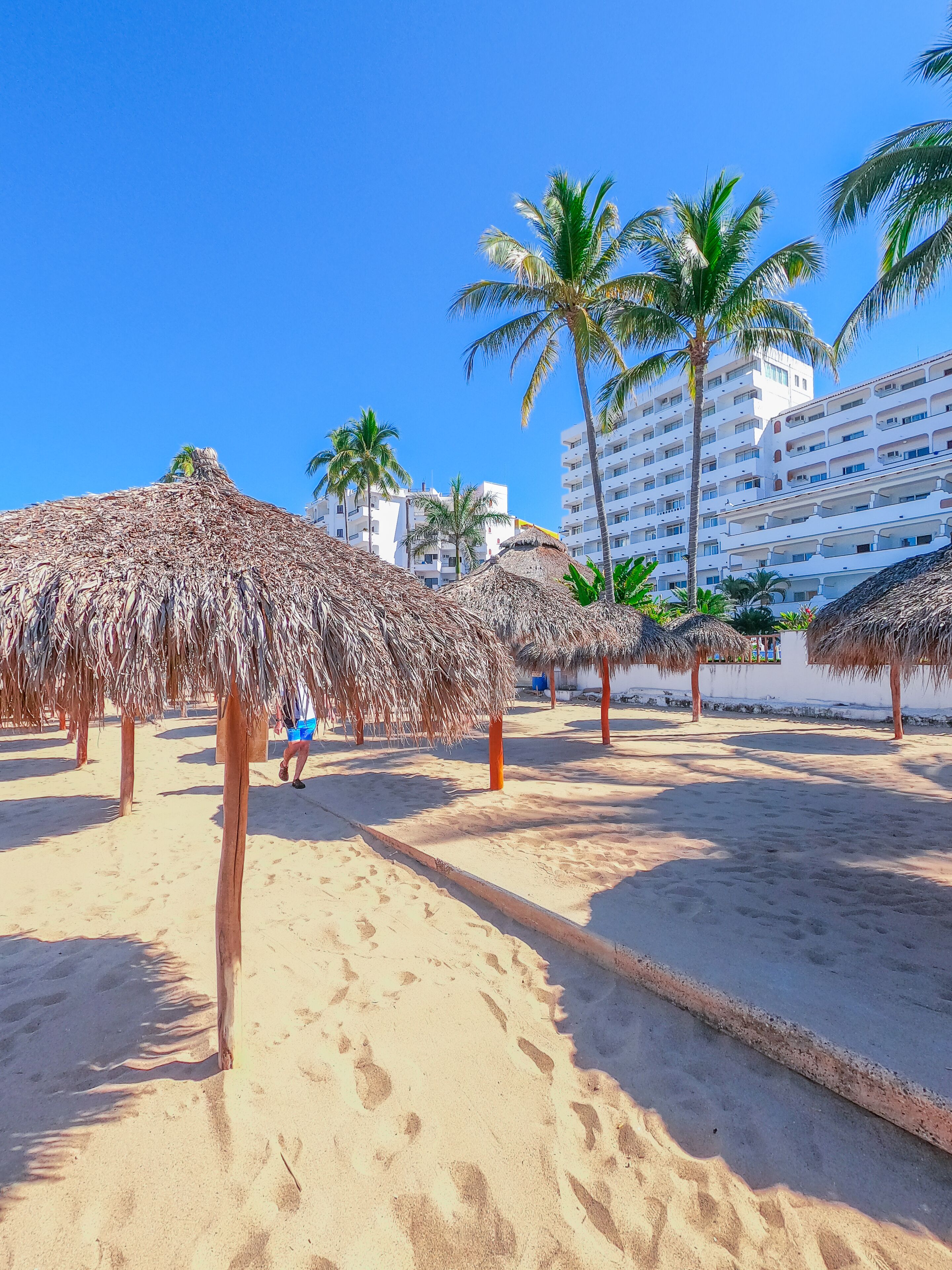 Een strand met rieten parasols, palmbomen en een hotel op de achtergrond.