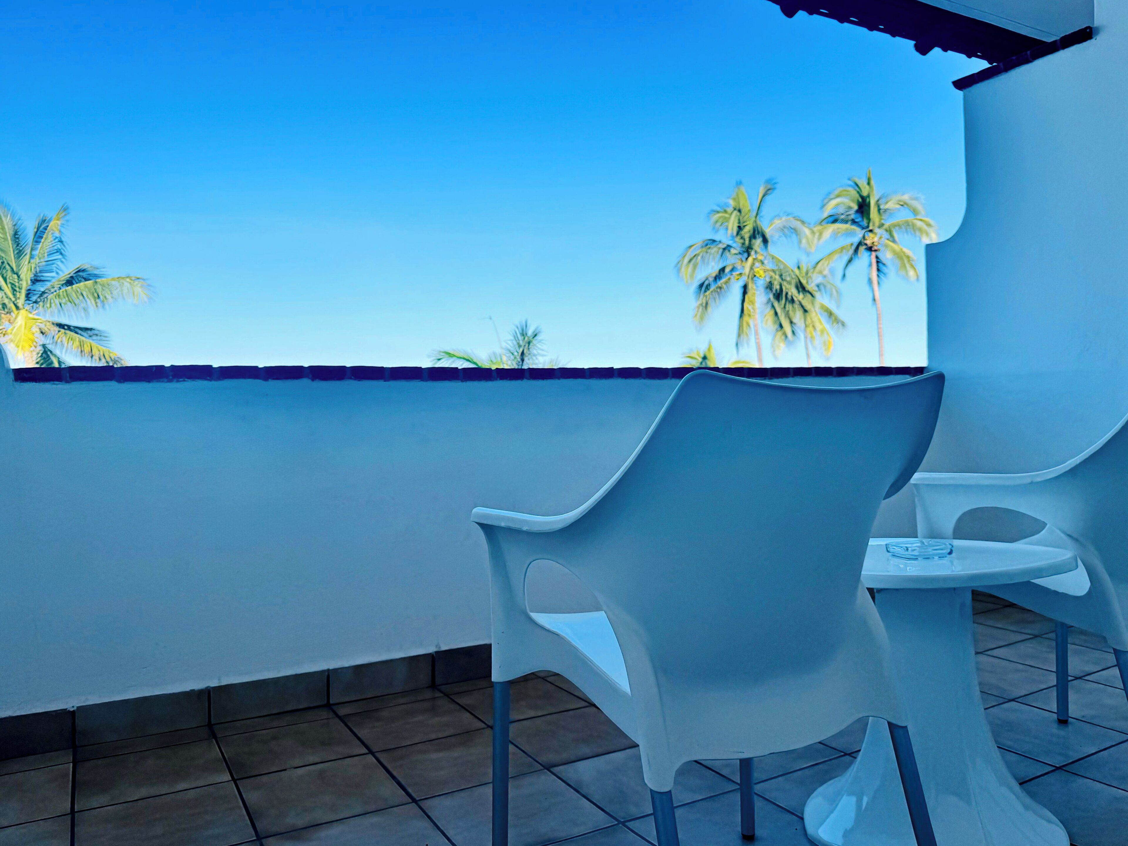 Un balcon avec deux chaises en plastique blanches et une petite table, d’où l’on peut admirer des palmiers et un ciel bleu dégagé.