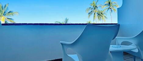 Un balcón con dos sillas de plástico blanco y una mesa pequeña, con vista a palmeras y un cielo azul despejado.