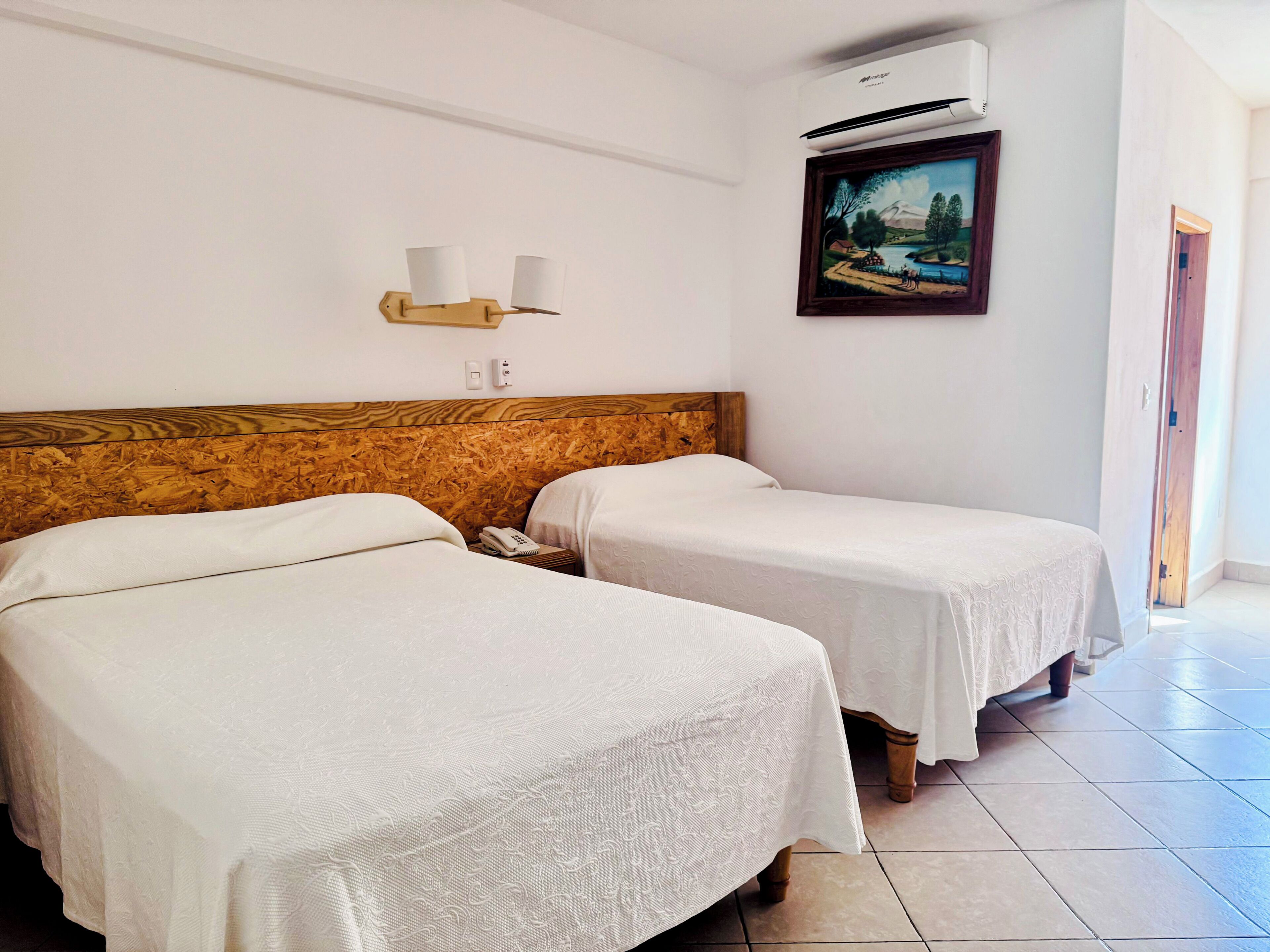 Une chambre d’hôtel avec deux lits, une tête de lit en bois, un tableau au mur et un appareil de climatisation.