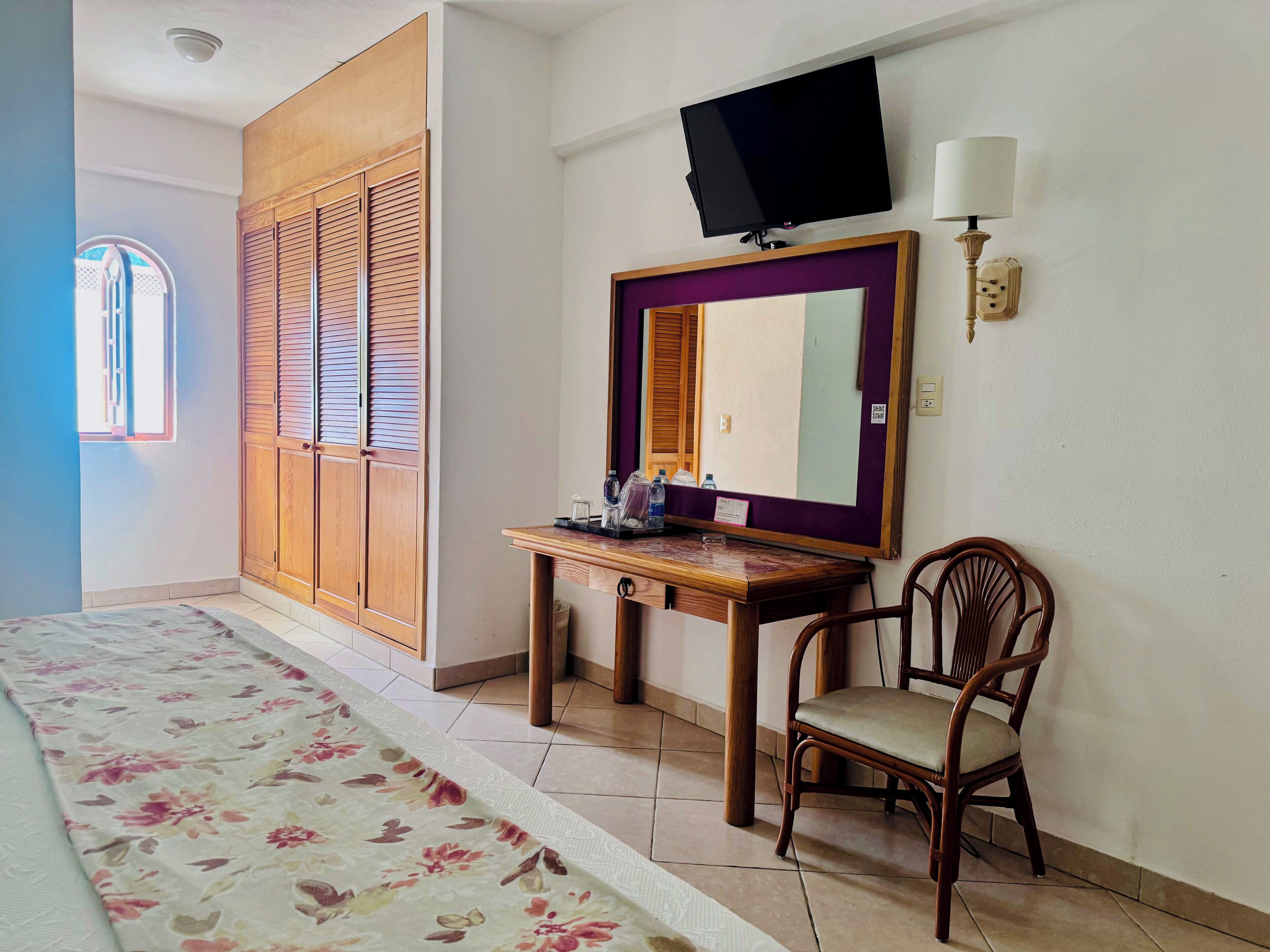A hotel room with a bed, a wooden desk, a chair, a television, and a window with shutters.