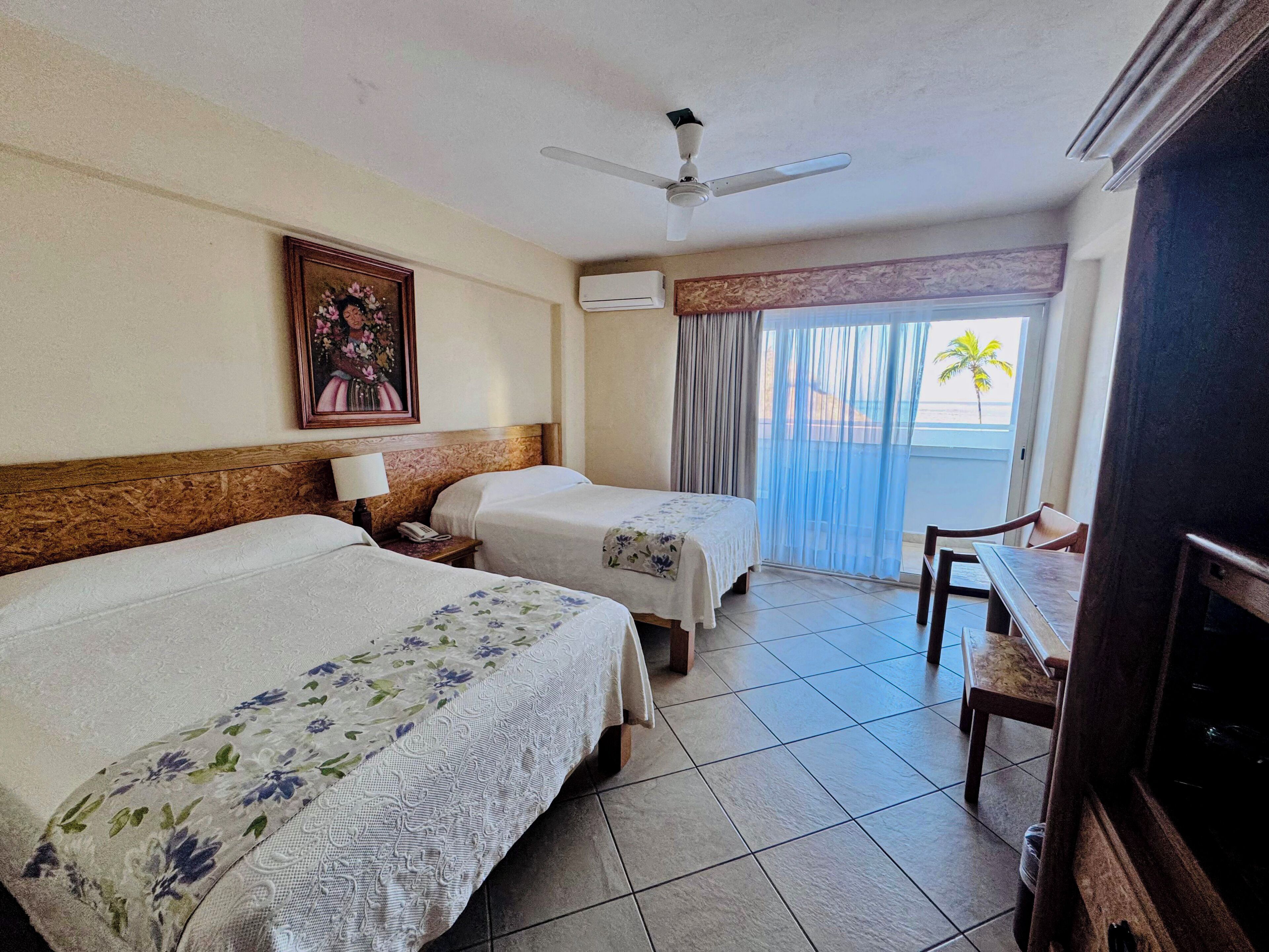 A hotel room with two beds, a ceiling fan, an air conditioning unit, a dining table with chairs, and a view of the outdoors.