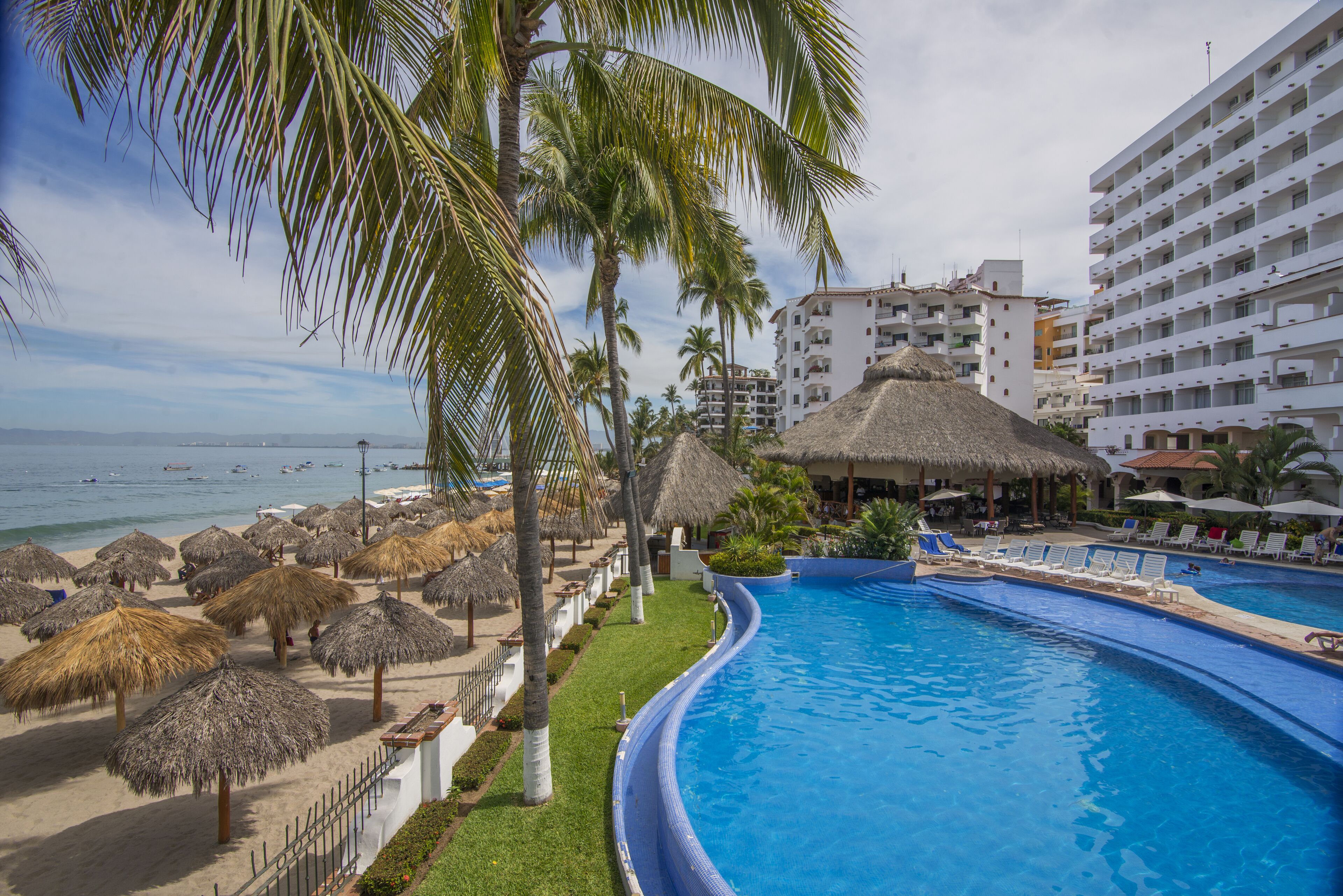 Zona de piscina en un resort con piscina curvo, palmeras y una playa con sombrillas de paja.