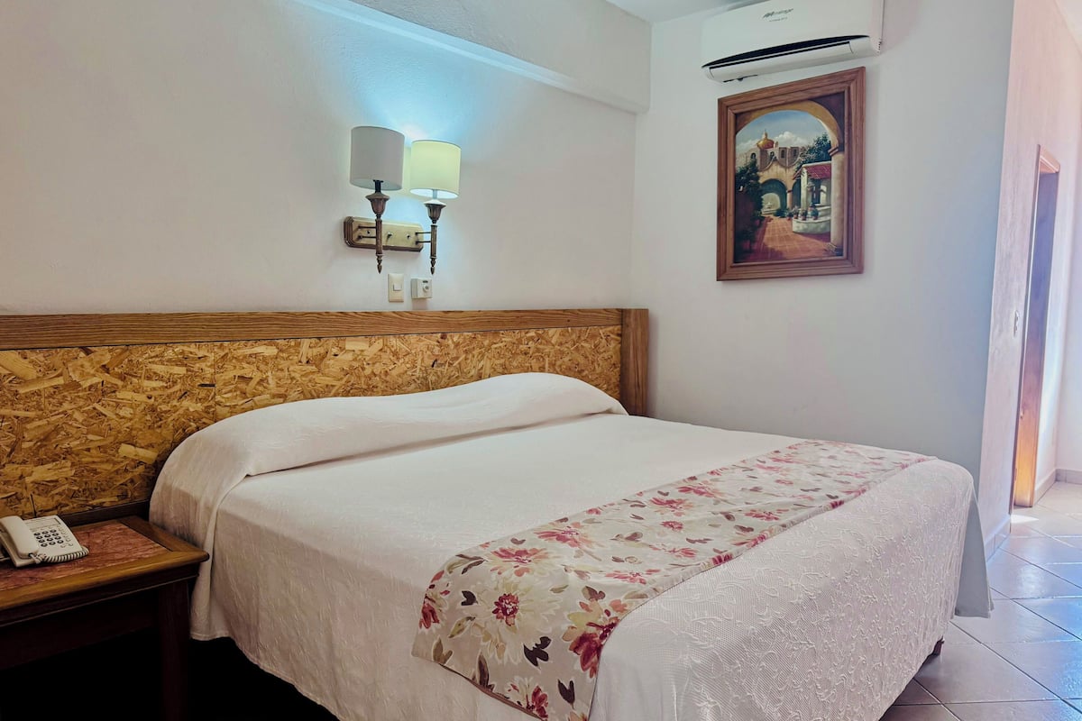 Una habitación de hotel con cabecera de madera, una cama con colcha estampada, una mesita de noche con teléfono, una lámpara de pared, aire acondicionado y un cuadro en la pared.
