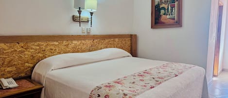 Una habitación de hotel con cabecera de madera, una cama con colcha estampada, una mesita de noche con teléfono, una lámpara de pared, aire acondicionado y un cuadro en la pared.