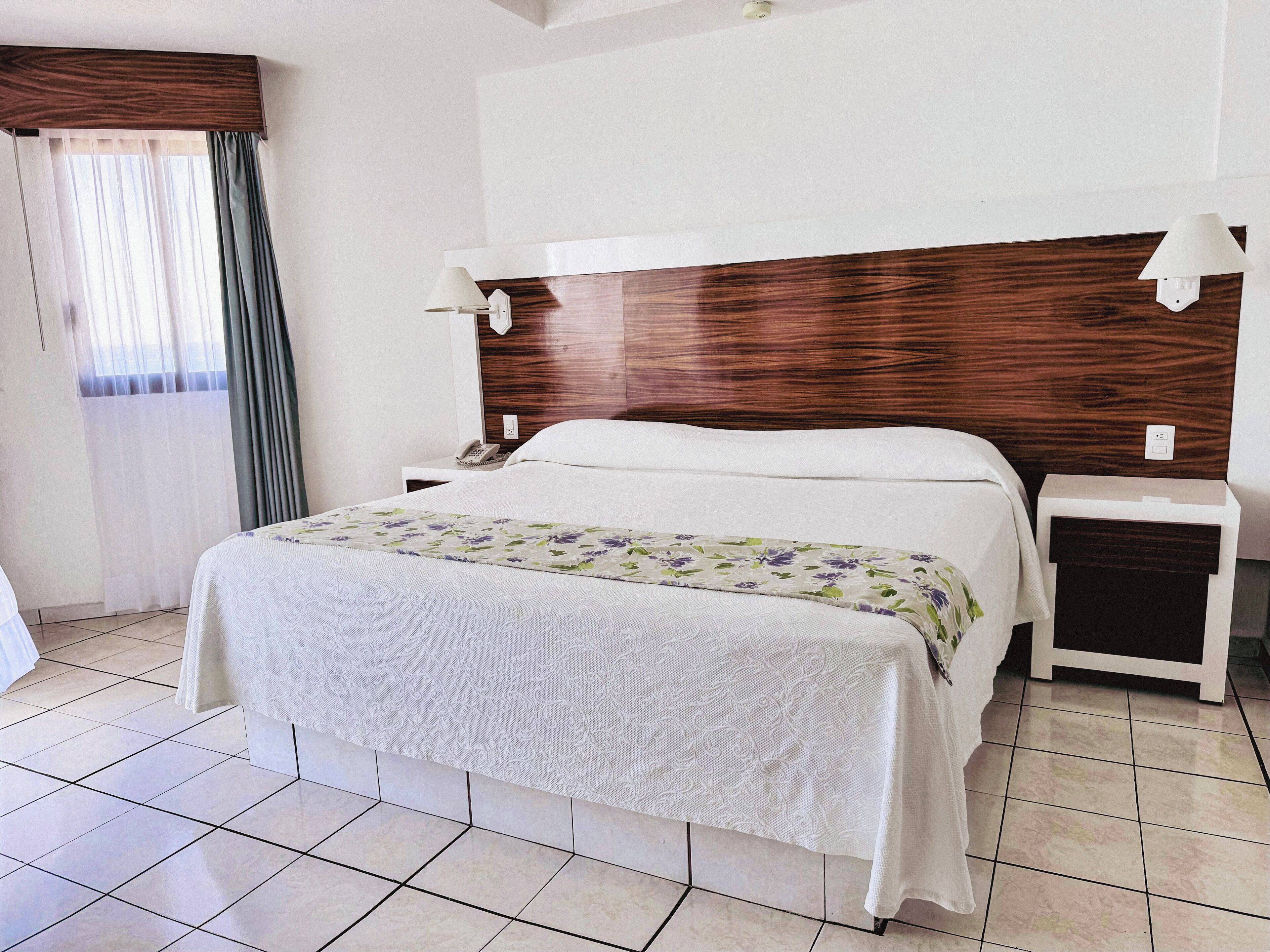 Quarto de hotel com uma cama, mesas de cabeceira, telefone e janela com cortinas.