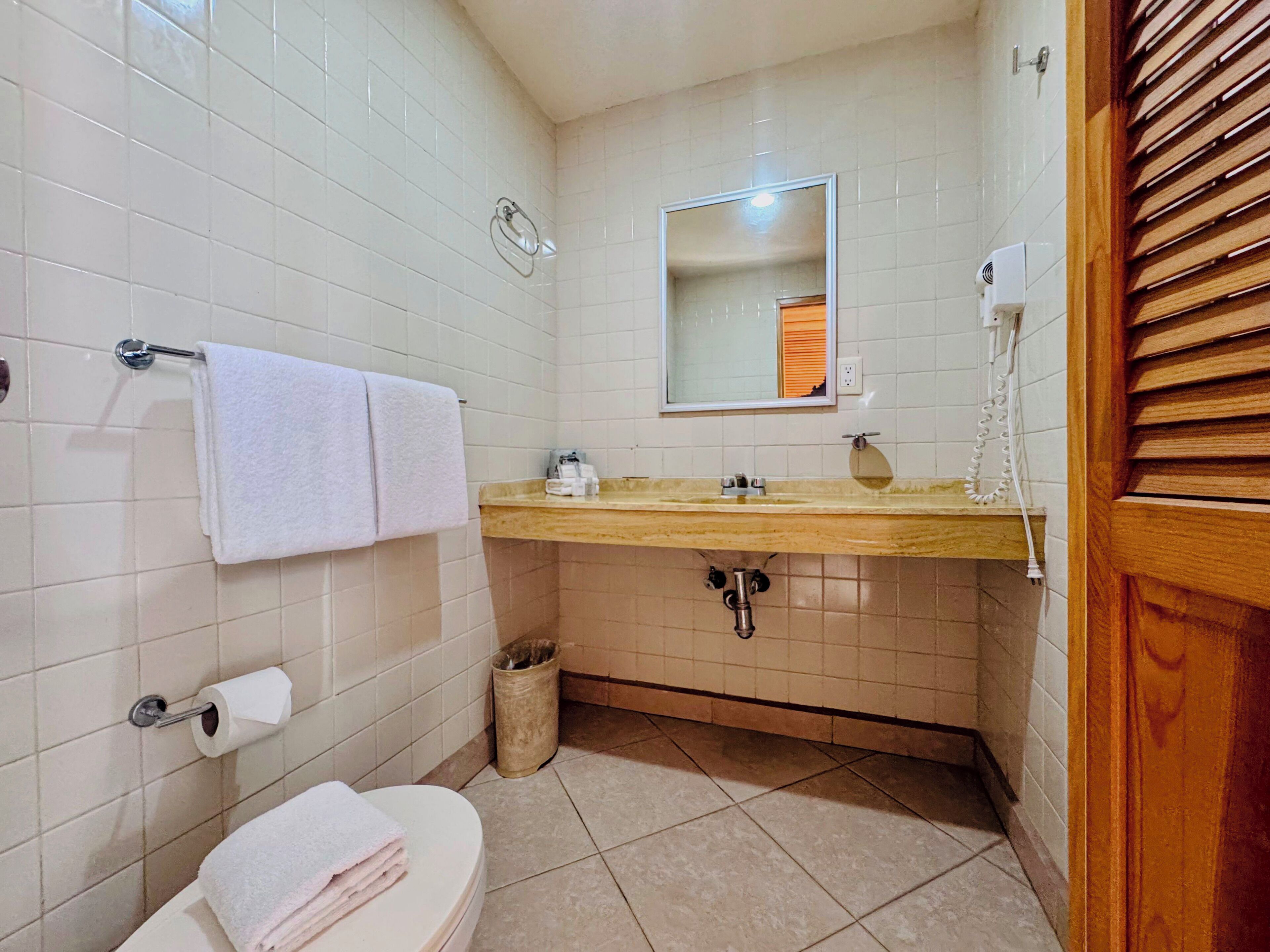 Une salle de bain avec des carreaux blancs, un meuble-lavabo en bois, des toilettes, un lavabo avec un miroir et un porte-serviettes.